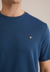 Laivyno mėlynos spalvos medvilninė marškinėlė su apvaliu iškirpimu ir trumpomis rankovėmis. Ant krūtinės yra mažas baltas ir juodas siuvinėjamas paukščio logotipas.