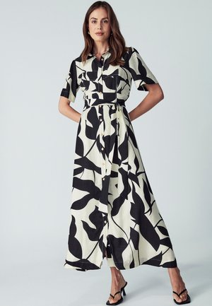 Kurzärmliges, cremefarbenes Hemdblusenkleid mit schwarzen Blattmustern, tailliertem Bündchen, fließendem Rock und seitlichem Schlitz. Getragen mit schwarzen High Heels.