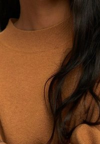 Brązowy sweter dzianinowy z prążkowanym dekoltem; miękka faktura; długie, faliste czarne włosy modelki opadają na ramię.