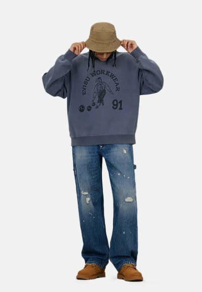 Felpa grigia con stampa grafica, abbinata a jeans blu strappati. Un cappello a secchiello beige e calzature marroni completano l'outfit.