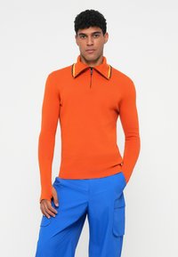 Suéter laranja de mangas compridas com colarinho de meia zip e riscas contrastantes amarelas e pretas, combinado com calças de carga azuis.