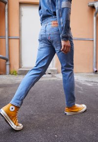 Džínové kalhoty ve světle modré barvě se slim fit a bledým vzhledem, doplněné o jasně žluté tenisky s vysokým límcem a černými akcenty.