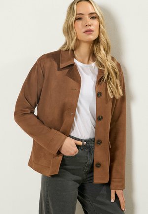 Blonde vrouw die een bruine jasje met knopen draagt over een wit shirt en zwarte spijkerbroek, staand met één hand in de zak tegen een effen muur.