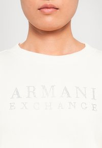 Detailní záběr na osobu, která má na sobě bílé tričko s malými lesklými nýty poskládanými napříč hrudníkem s nápisem "ARMANI EXCHANGE".