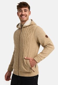 Sudadera de punto beige con capucha forrada de sherpa, cierre frontal, dos bolsillos laterales y un parche de logo marrón en la manga izquierda.