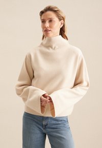 Maglione girocollo beige chiaro in stoffa morbida e texturizzata con maniche ampie e svasate. Abbinato a jeans in denim blu per creare contrasto.