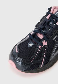 Baskets de sport noires avec des accents roses, tige en maille, semelle texturée et détails marqués. Comprend un laçage traditionnel et un capuchon renforcé.