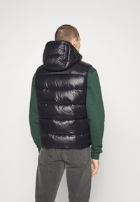 Schwarze gesteppte Weste mit Kapuze, glänzender Textur und steppendem Design. Wird über einem grünen Sweatshirt getragen, darunter sind graue Jeans sichtbar.