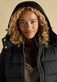 Noor blondide lokkidega noor naine kannab musta kapuutsiga sulejaki ja mustriga särki, vaatab otse ja tal on kerge naeratus näol.