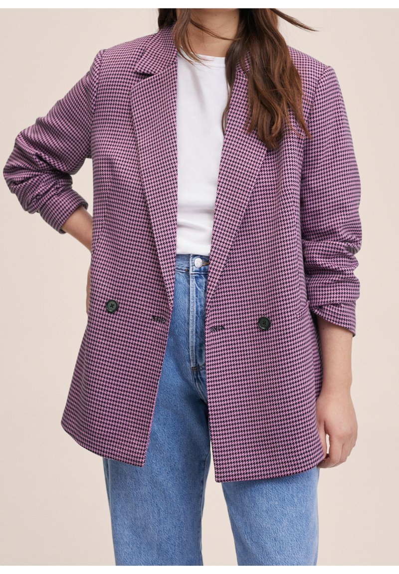 Femme portant un blazer violet et noir à motif pied-de-poule sur une chemise blanche et un jean bleu, une main sur la hanche et de longs cheveux bruns visibles.