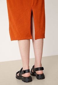 Pomarańczowa sukienka midi z rozcięciem z tyłu, zestawiona z czarnymi sandałami z grubą podeszwą i regulowanymi paskami. Gładka faktura materiału.