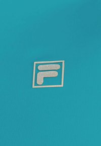 Tejido verde azulado con una textura acanalada que presenta un emblema dorado y brillante en un contorno cuadrado, mostrando un diseño estilizado de la letra 'F'.