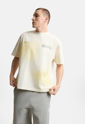 Młody mężczyzna ubrany w luźną, kremową koszulkę z subtelnymi żółtymi plamami oraz szare spodnie dresowe, patrzący bokiem na białym tle.