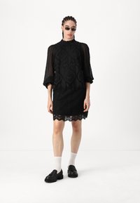 Sort kjole med et teksturert mønster, puffer transparente ermer og bølget kant. Kombinert med svarte sko og hvite sokker.
