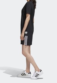 Kvinna iklädd en svart Adidas T-shirtklänning med vita ränder och vita Adidas sneakers med svarta ränder, ståendes i profil.