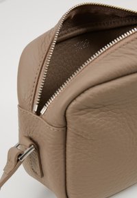 Béžová kožená taška s texturovaným povrchem, čtvercového tvaru a stříbrným zipem po horním okraji. Obsahuje boční úchyt.