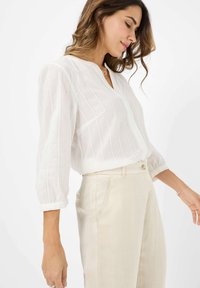 Camicia bianca, leggera e trasparente, con righe verticali, scollatura a V e maniche a tre quarti, abbinata a pantaloni beige chiaro.
