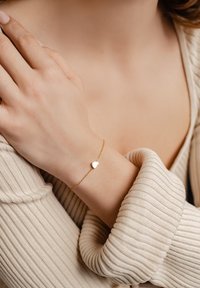 Pulsera de cadena de oro con un pequeño charm en forma de corazón. Lleva en una muñeca, mostrando su textura suave y diseño delicado.