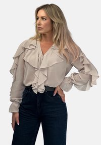 Jasno beżowa bluzka z falbankami z przodu i rękawami, wykonana z przezroczystego materiału, zestawiona z ciemnymi dżinsami o wysokim stanie. Posiada dekolt w kształcie litery V.