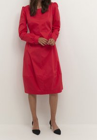 Rode jurk met lange mouwen en een vierkante halslijn, geplooide schouders en gathered mouwen. Gladde, lichte stof; knie-lang met een rechte snit.