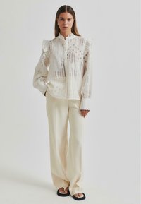 White, textured blouse with ruffled shoulders, featuring cut-out patterns; paired with cream trousers and black flip flops.