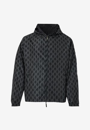 Mand iført en sort mønstret hættetrøje, lyseblå t-shirt, slidte løse blå jeans og hvide sneakers, stående mod en ensfarvet baggrund.
