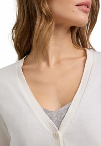 Witte cardigan met een V-hals en knoopsluiting. Kenmerkt zich door textuursteken langs de randen en is over een grijze top gedragen.