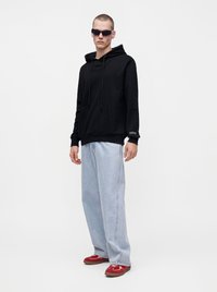 Homme portant un sweat à capuche noir, un jean large bleu clair, des baskets rouges et des lunettes de soleil foncées, debout sur un fond blanc uni.
