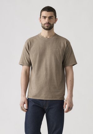 Moški z brado in kratkimi lasmi, oblečen v rjavo Levi's majico in temno modre kavbojke, stoji pred enobarvnim svetlim ozadjem.