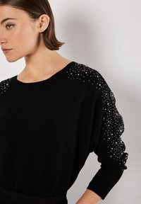 Schwarzer Pullover mit lockerem Schnitt, strukturiertem Strick, verziert mit silbernen Perlen an den Schultern und Ärmeln. Hat einen weiten Ausschnitt und lange Ärmel.