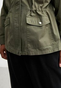 Olivgrüne Jacke mit verstellbarem Bund, zwei Vordertaschen, Druckknöpfen und einer glatten, matten Textur. Beinhaltet geraffte Details.