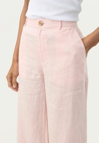 Helle rosa Leinenhosen mit lockerer Passform, ausgestattet mit einem einzelnen Holzknopf und seitlichen Taschen. Die Textur ist leicht zerknittert.