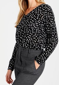 Blouse noire à motif d'étoiles blanches, col en V, manches longues, et tissu fluide ; assortie à un pantalon gris à motifs taille haute.