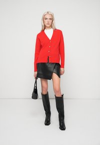 Cardigã vermelho com decote em V e cinco botões, combinado com uma minissaia preta de couro. Botas pretas até ao joelho e uma pequena carteira completam o visual.