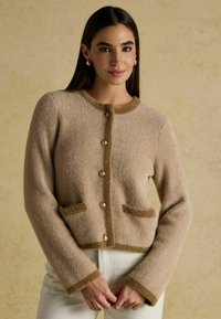Bež herringbone musteriga kardigan pehme pruunide äärtega, mis on varustatud kuldsete nööpide ja kahe esiseguga taskuga; tekstureeritud kudum, lühendatud disain.