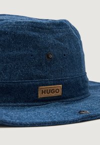 Blå denim bøttehatt med bred skjerm, med et brunt merkelapp og svarte metalldetaljer på sidene. Synlige sømmer.
