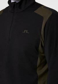 Närbild av en svart zip-up jacka med olivgröna paneler på axlarna och sidorna, med en liten minimalistisk logotyp på bröstet.