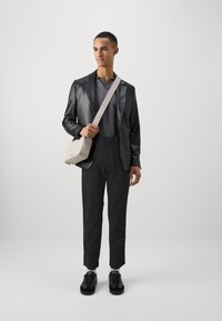 Blazer in pelle nera sopra un polo testurizzato grigio scuro, abbinato a pantaloni neri e sneaker nere. Borsa a tracolla beige con tracolla regolabile.