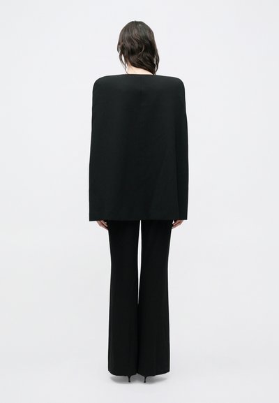 Pikkade tumedate juustega naine seisab seljaga kaamera poole, kandes valgel taustal struktuurset musta laiade õlgadega jopet ja põlvpükse.