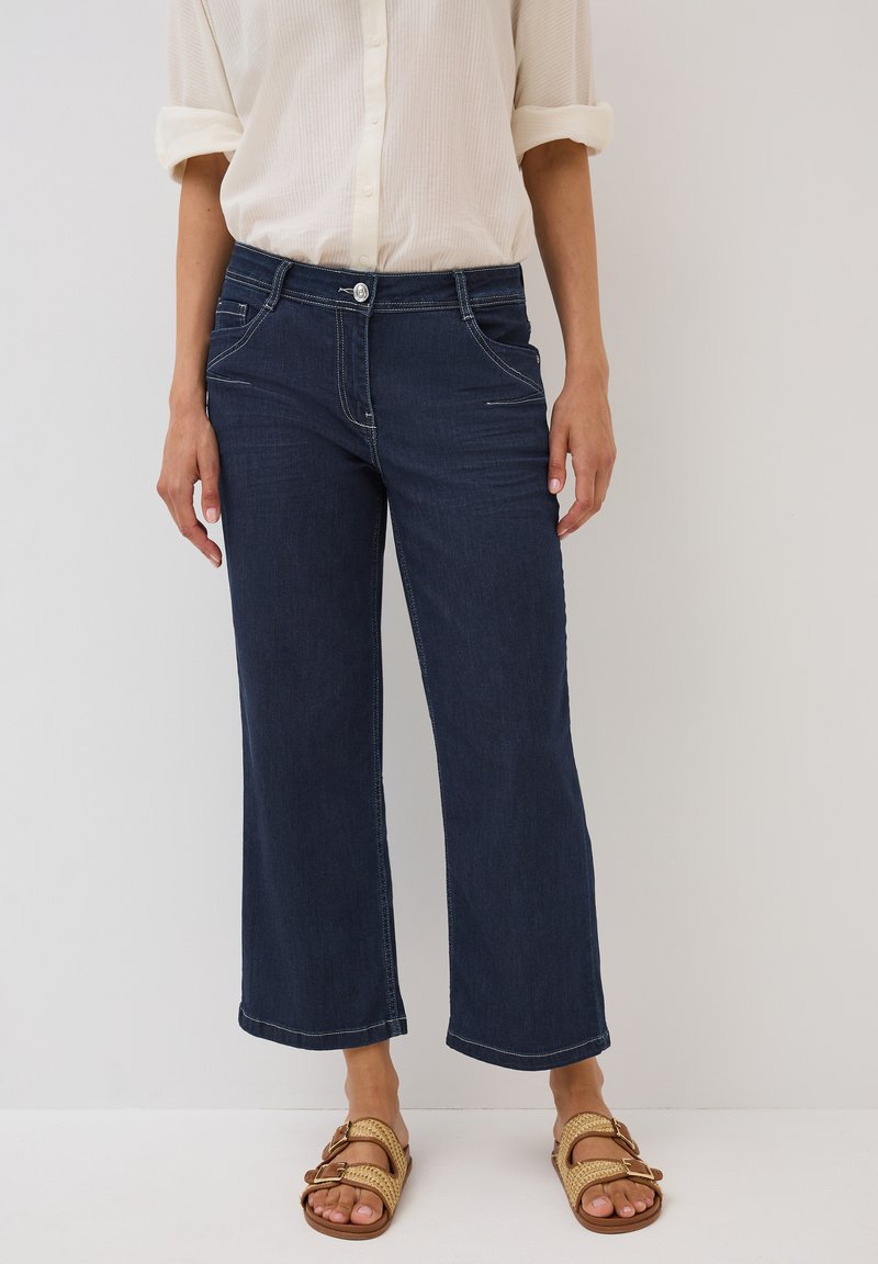 Jeans a gamba larga di un blu scuro con vita media, dotati di tasche frontali, cuciture chiare e chiusura con bottone argento. Indossati con sandali beige.
