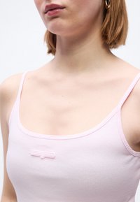 Femme aux cheveux courts châtain clair portant une boucle d'oreille en or en forme de créole et un débardeur côtelé rose clair avec un petit logo sur la poitrine.