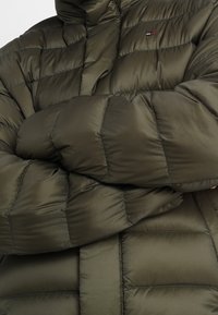 Olivgrön puffjacka med kviltat mönster, som har en hög krage och broderad logotyp. Materialet verkar glänsande och isolerat.