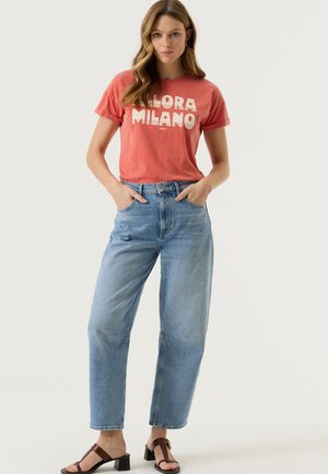 Donna che indossa una maglietta rossa "Allora Milano", jeans a vita alta azzurri chiaro e sandali marroni con tacco, in piedi con le mani nelle tasche.