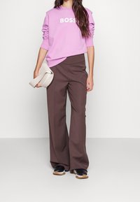 Růžová mikina s logem "BOSS", hnědé kalhoty s vysokým pasem a širokým střihem, a černé tenisky s béžovou podešví. Drží béžovou tašku.