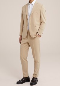 Beige pak met een getsailored blazer en broek, gecombineerd met een wit overhemd. Gladde textuur; minimale details; gedragen met donkere loafers.
