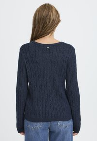 Námornícky modrý sveter s vrkôčkovým vzorom, dlhými rukávmi, okrúhlym výstrihom a ribovaným lemom. Mäkka textúra s jemným leskom; obsahuje malý štítok značky.