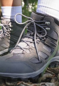Gray hiking boots with mesh and suede panels, featuring metal eyelets and laces. Positioned on a natural surface with greenery in the background.