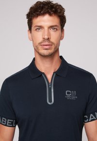 Granatowa koszulka polo z zamkiem przy kołnierzu, szarymi akcentami oraz logo na klatce piersiowej i rękawie. Wykonana z teksturowanej dzianiny.