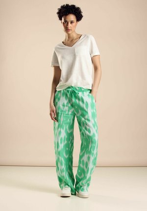 Locker sitzende grüne Hosen mit einem Batikmuster, aus leichtem Stoff gefertigt, kombiniert mit einem weißen V-Ausschnitt-Shirt und weißen Sneakers.