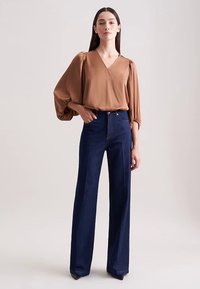 Braune Bluse mit V-Ausschnitt und Puffärmeln, kombiniert mit hoch taillierter, weit geschnittener dunkler Jeans. Glatter Stoff und klare Linien.